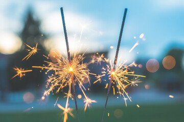 A close-up photograph of two golden sparklers burning against a blurred background.