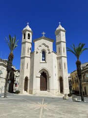 La facciata in stile stile gotico-moresco della chiesa del ss. Crocifisso a Rosolini in pietra bianca su cielo azzurro. Due palme fanno da cornice.