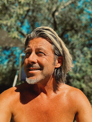 Smiling man with blond half long hair and grey stubble beard in sunny garden by an olive tree.