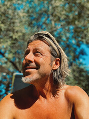 Smiling man with blond half long hair and grey stubble beard in sunny garden by an olive tree.