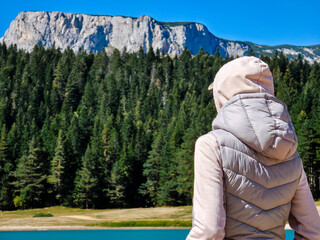 Person facing the mountains and forest by Black Lake in Durmitor National Park. A calm moment of solitude and connection with nature, framed by blue sky, pine trees and rugged rocky peaks.