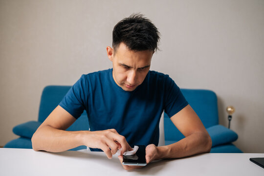 Portrait of focused young man cleaning screen of smartphone with disinfectant wipe, promoting hygiene and effectively eliminating germs and bacteria for better health, sitting at white home table.