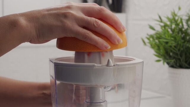 A hand presses half a grapefruit onto the plastic reamer of an electric citrus juicer. The motor engages with a loud whirring sound as the fruit is squeezed to extract the juice.