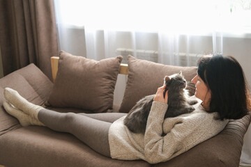 Smiling woman is lying on brown sofa, stroking at fluffy gray cat resting on her chest, bathed in window sunlight. Pet companionship, relaxation, cozy home life, love for animals
