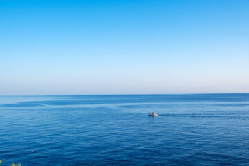 Small fishing vessel upon calm blue sea under clear sky, viewed from distance within soft daylight