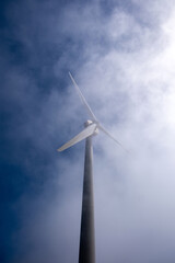 Wind turbine tower rising into hazy sky, blades cutting through soft mist and bright daylight image