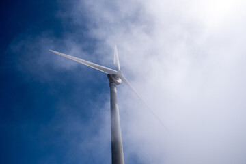 Wind turbine tower rising through bright morning mist, blades cutting into clear blue sky image