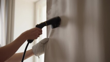 Homemaker using manual garment steamer to clean and refresh curtains with hot vapor, effectively removing wrinkles, dust mites and bacteria for hygienic home environment, close-up, slow motion.