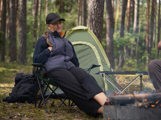 Female camper relaxing in a folding chair beside a campfire and tent, savoring a tranquil moment in the serene woods during an enjoyable camping trip in nature