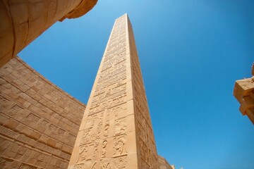 Karnak Temple complex obelisk piercing the blue sky, intricate hieroglyphs visible. A close up, low angle shot of a towering granite obelisk at Karnak Temple in Egypt, reaching towards a clear, deep