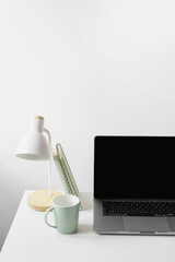 A close-up workspace shot with a laptop, mint mug, white desk lamp, and green notebook in a minimal and bright setting.