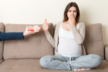 Pregnant woman sitting on the sofa rejects to eat junk food such as donuts and makes no gesture. Healthy diet for future mother concept