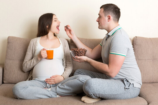 Happy husband is feeding his pregnant wife with chocolate cereal balls and juice relaxing on the sofa at home. Future father takes care about expecting mother