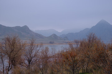 A cold misty landscape and the outlines of mountains covered with haze, against the background of the calm surface of the Ili river, framed by leafless autumn vegetation, Kazakhstan.