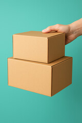A person's hand holds two stacked plain brown cardboard boxes against a solid turquoise background.