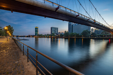 A tranquil dusk scene along the river featuring a large suspension bridge overhead, warm street lights, and modern office towers reflecting on calm water for urban lifestyle and architecture themes.