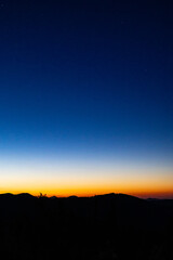 Predawn gradient from deep navy to warm orange rises above layered mountain silhouettes. Minimal, vertical sky scene with faint stars, clean copy space, and calm atmosphere ideal for backgrounds.