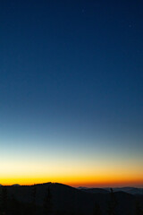 Golden band of sunrise edges the horizon beneath a vast blue sky, revealing layered mountain ridges in silhouette. Vertical minimal landscape with faint stars, crisp air, and generous copy space.