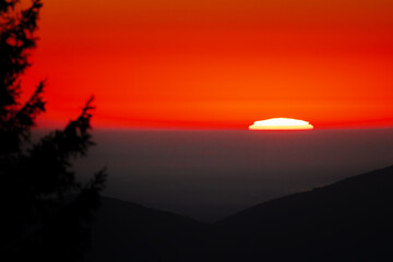 Fiery sunset dips behind layered mountain ridges, casting a radiant gradient from deep crimson to...