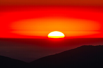 Close view of the glowing sun touching the horizon, in intense red and orange halos above shadowy ridges. Dramatic light and color create an evocative background for travel, wellness, and ads UX.