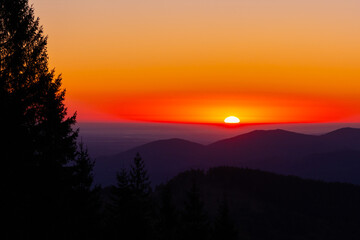 Sunset bathes layered mountains in the vivid orange red glow. Dark spruce silhouettes edge the frame, adding depth and calm. Perfect for travel ads wellness brand, prints, and calm web banners.