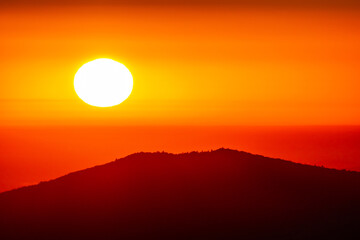 Fototapeta premium Sunset bathes layered mountains in the vivid orange red glow. Dark spruce silhouettes edge the frame, adding depth and calm. Perfect for travel ads wellness brand, prints, and calm web banners.