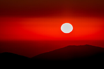 Obraz premium Intense crimson sunset with the sun as a bold white disk above a dark mountain ridge. Minimal, dramatic, and moody, this vivid backdrop suits posters, websites, wellness themes, and travel media.