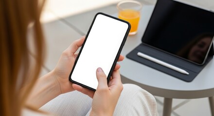 Woman using a smartphone with a blank white screen next to a tablet and orange juice on a table perfect for showcasing app designs or mobile content ideas