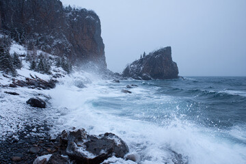 Rocky coastline during winter storm, icy waves crashing, snow-covered cliffs, cold dramatic atmosphere, realistic texture details