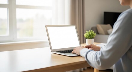 Unrecognizable person typing on a laptop computer with a blank white screen, working from a home office desk with natural light