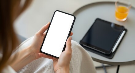 Woman holding a blank screen smartphone with a tablet and orange juice on a table in a bright modern interior providing a mockup for app or website design
