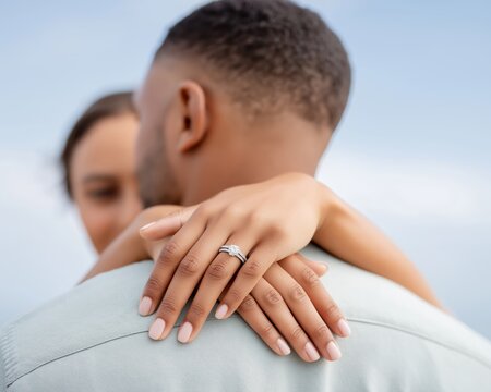 Romantic moment outdoors with a close-up of a woman's hands embracing her partner, highlighting a sparkling diamond engagement ring against a soft blue sky background