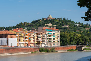Scenic view of Verona, Italy, with buildings lining the Adige River and a lush, green hillside in the background with the Santuario della Madonna di Lourdes.
