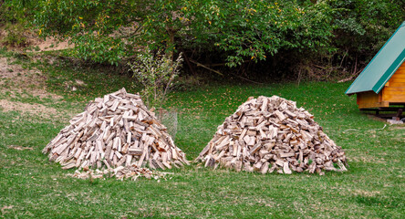 Two large piles of chopped firewood stacked on green grass in a rural backyard. Dry split logs prepared for winter heating. Natural rustic scenery with trees and autumn leaves in the background.
