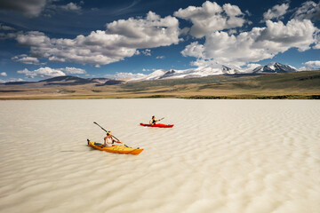 Two female tourists are walking on kayaks at white water mountain lake against snow capped mount at bright sunny day. Active vacations concept