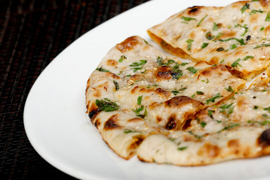 A close-up view of freshly baked, soft Naan or Kulcha, an Indian flatbread, showing its charred spots, cilantro garnish, and glossy buttered surface on a white restaurant plate.