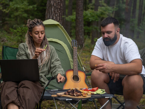 Young woman freelancer working on a laptop while enjoying grilled food at a campsite in the woods, with a tent and guitar nearby, embracing the tranquility of nature - Powered by Adobe
