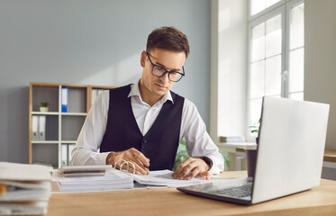 Fototapeta premium Portrait of young business man accountant in suit and glasses working at the desk with a pile of documents on his workplace in office. Company employee sitting with financial documents and laptop.