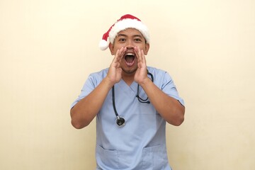 Southeast Asian male nurse in Santa hat and scrubs shouting or making announcement, isolated on plain background, ideal for festive ads.