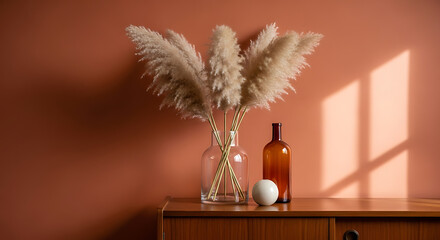 Pampas grass in a clear glass vase with brown bottle and white sphere on a wood cabinet against a terra cotta wall with window light.