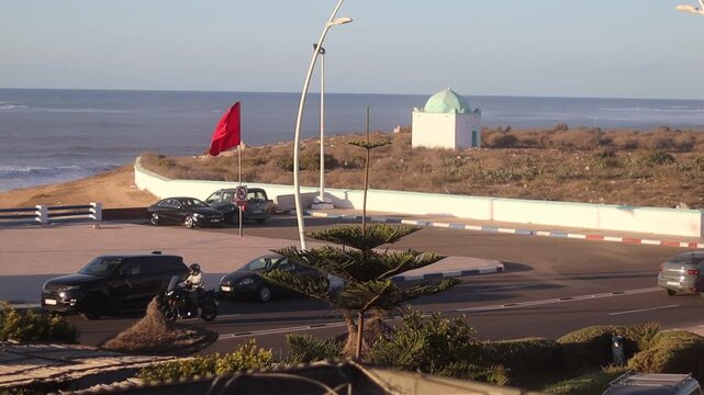 Coastal roundabout traffic with waving Moroccan flag and white domed shrine in Sidi Bouzid, Morocco, cars turning at oceanfront circle, cultural landmark