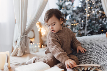 Little boy sitting on the bed in front of christmas tree. Toddler playing with toys and candles. Brown colors.