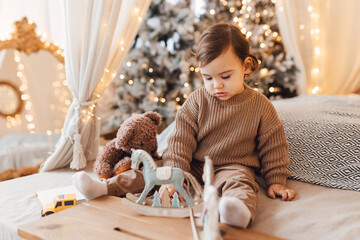 Little boy sitting on the bed in front of Christmas tree. Toddler playing with toys