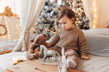 Little boy sitting on the bed in front of Christmas tree. Toddler playing with toys