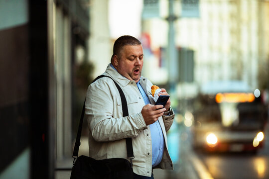 Adult man surprised by phone while eating pastry at bus stop - Powered by Adobe