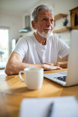 Senior man using laptop at kitchen table, thoughtful