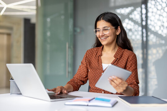 Young smiling indian woman multitasking, using a laptop and digital tablet for work, showing happiness and productivity in a modern office environment
