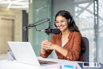 Young woman smiling while recording an audio podcast using a professional microphone, headphones, and laptop for online streaming content from a modern studio or office