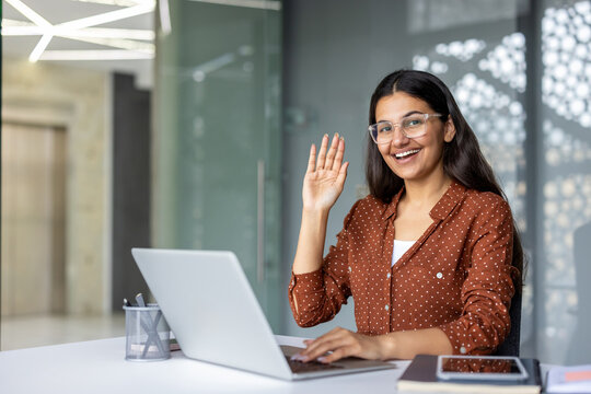 Indian woman in a bright home office smiling and waving during a laptop video call, conveying remote work, virtual communication, professionalism and friendly greeting