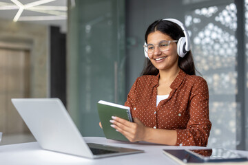 Young woman student wearing headphones smiles, holding a notebook while participating in an online...